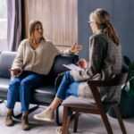 A counselor sits in a wooden chair, taking notes, while her client, another woman, engages in expressive conversation with both hands gesturing in the air