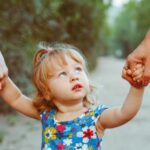 A toddler takes cautious steps, glancing upward at an unseen parent whose hand provides a reassuring anchor. Another young child gently places their hand on the toddler's back