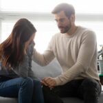 a male and female couple holding hands on a couch. The female is crying, the male consoles her.