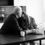 elderly father consoling daughter. Both sit at a table, the daughter leans into her father as he puts his arm around her