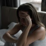 A woman sits alone on her bed, visibly distressed, holding the side of her head.