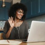 woman on laptop in kitchen on video call, waves at her screen and smiles