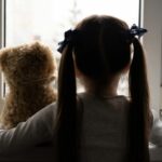 young girl puts her arm around her teddy bear as she looks out of a window