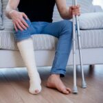 a person from the chest down, sitting on a couch with their leg in plaster. Their left hand is holding two crutches.