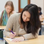 female student in plain clothes completes exam at a desk in exam hall