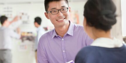 A man and a woman are having a conversation in in an office environment. The woman's back is to the camera and the man is smiling.