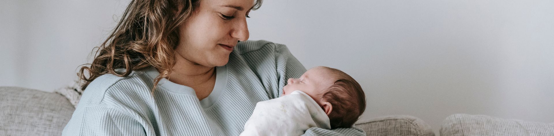 A mother sits on the couch while holding and looking down at her baby.