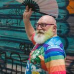 Man with rainbow t-shirt on, smiles over his shoulder towwards camera, whilst waving a fan above his head.