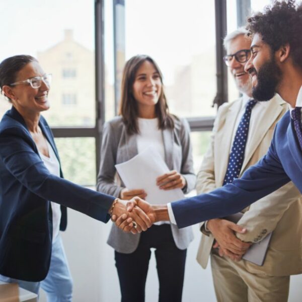 Two people shake hands smiling while standing with a small group of people