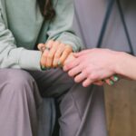 a woman can be seen sitting from the chest down. Another person is holding her hand in a supportive way.