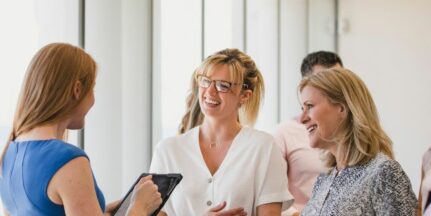 Three women stand in a bright office buidling holding books or tablet devices, smiling and talking
