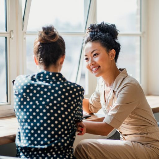 two women sitting in front of a window smiling and talking