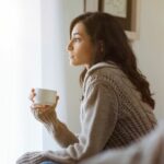 women sitting in chair, with a coffee cup in one hand, contemplatively looking out the window