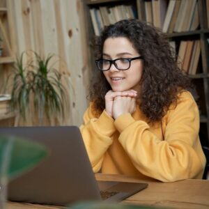 A woman sits at a desk with hands under her chin, paying attention to a laptop screen and smiling