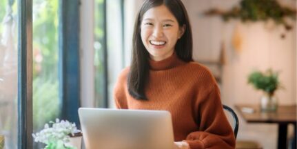 A woman sits in a bright home officer with her laptop, smiling at the camera