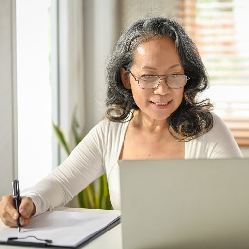 A woman sits in a home office working on her laptop and taking notes in a notebook.